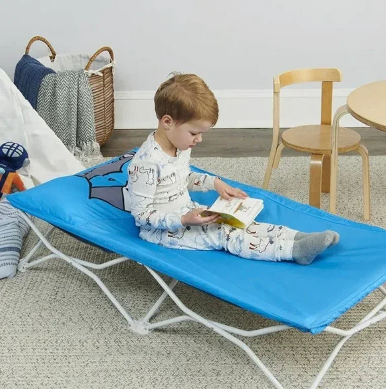 Young boy in pajamas reading a book on a blue folding kids cot in a playroom.