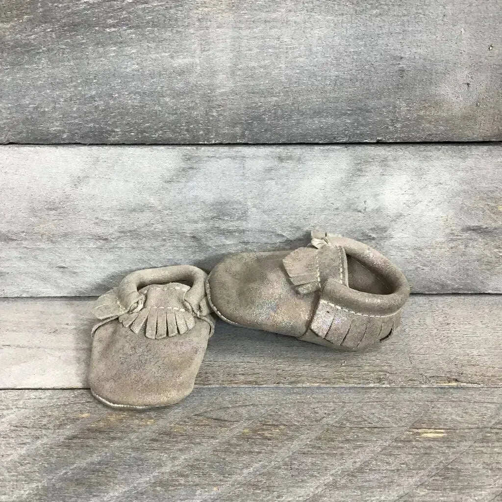Angled view of Freshly Picked soft sole leather moccasins in iridescent silver showcasing fringe trim on grey wood plank floor