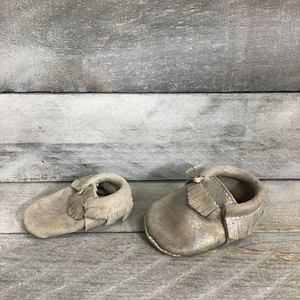 Front and side view of silver holographic leather baby moccasins showing fringe detail and soft soles on gray wood background