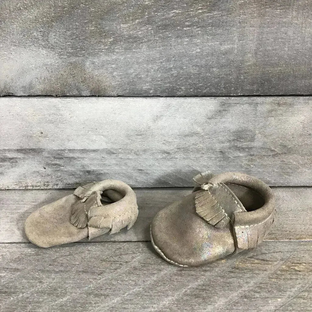 Front and side view of silver holographic leather baby moccasins showing fringe detail and soft soles on gray wood background
