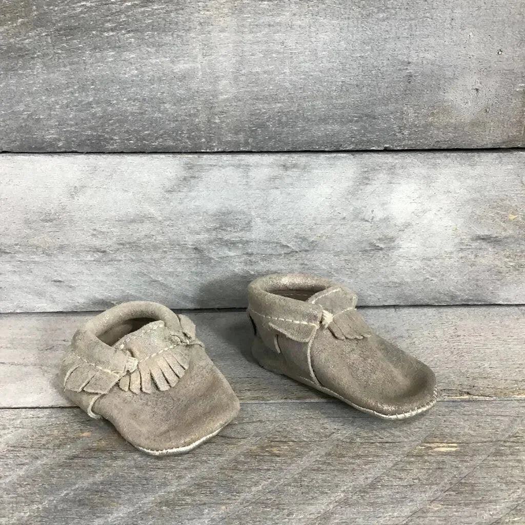 Three-quarter view of soft-sole leather baby moccasins showing fringe and iridescent silver finish on gray wood background