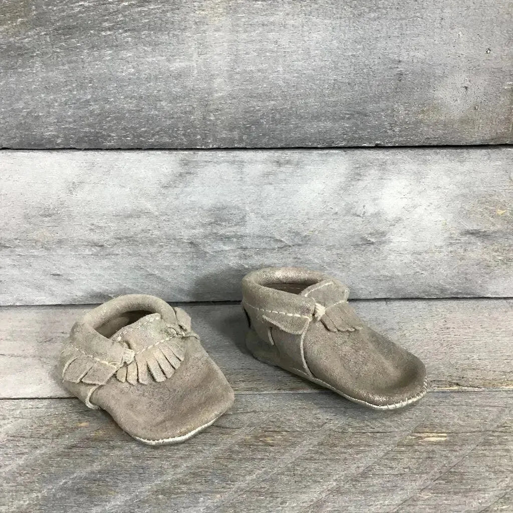 Three-quarter view of soft-sole leather baby moccasins showing fringe and iridescent silver finish on gray wood background