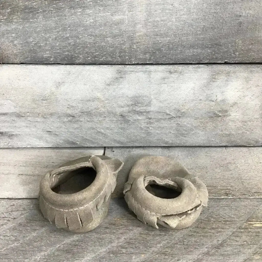 Angled top view of used silver Freshly Picked soft-sole leather moccasins showing fringe detail and scuffed soles on a gray wood background