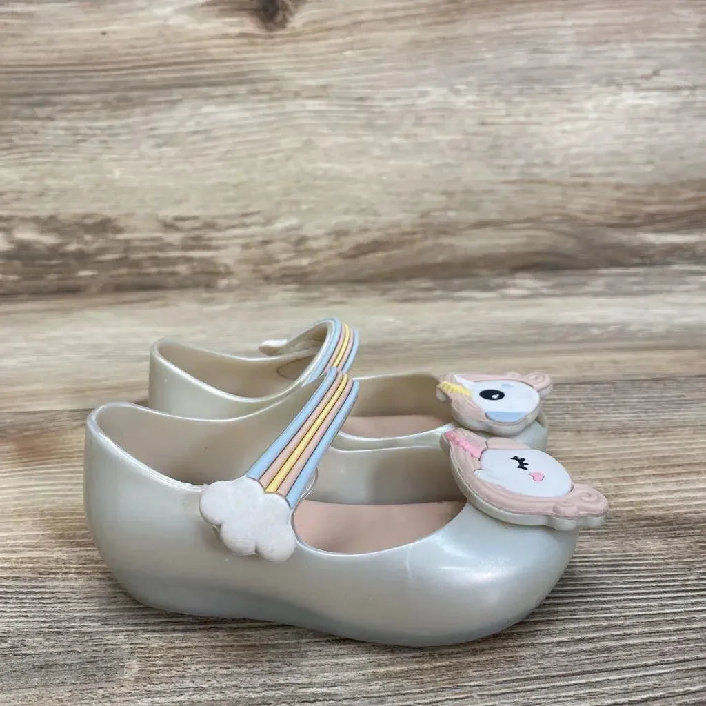 Side view of ivory Mini Melissa Glitz Unicorn Mary Jane toddler shoes showcasing unicorn face appliques and rainbow cloud straps on wood floor background
