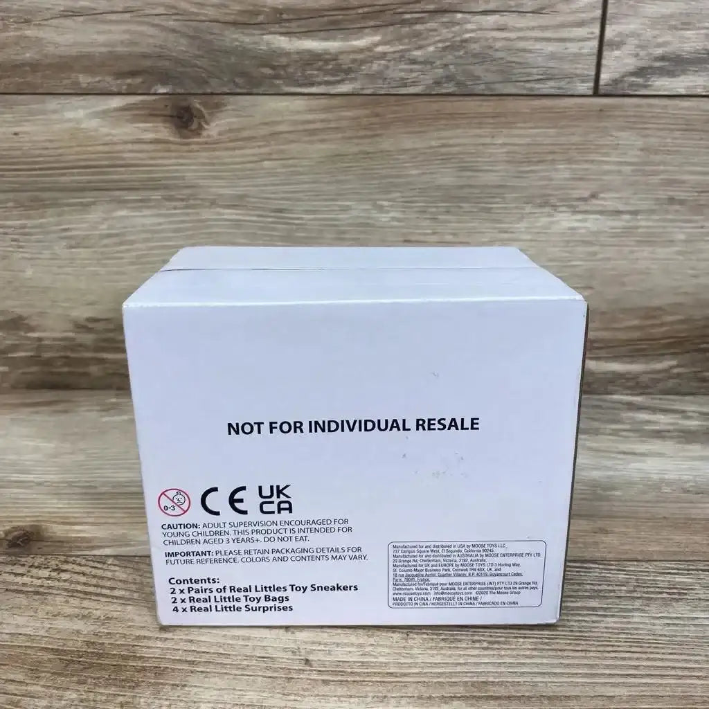 Back view of Real Littles sneaker and bag bundle box showing NOT FOR INDIVIDUAL RESALE headline, safety symbols, and contents list on wood tabletop