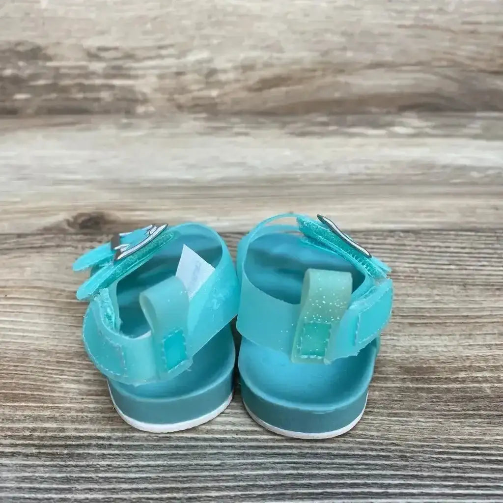 Back view of teal newborn jelly sandals showing glitter ankle straps, heart buckle, and Velcro on wood background