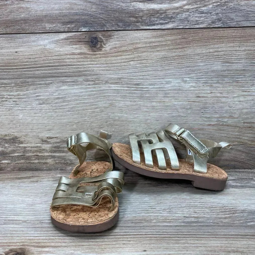 Three-quarter view of gold OshKosh Raquel toddler sandals with cork footbed, adjustable ankle strap, and cutout metallic upper on wood backdrop