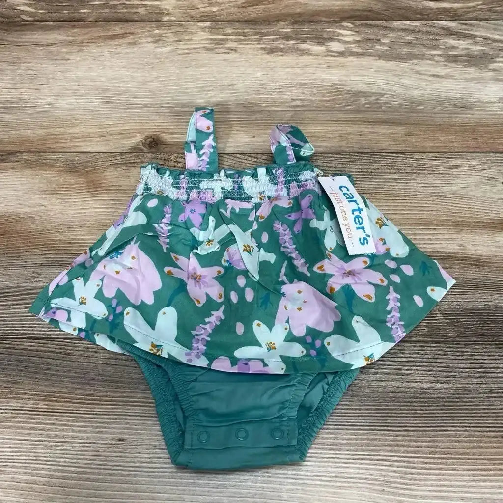 Front view of baby girl's green floral bodysuit dress showcasing ruffled skirt, shoulder straps, and three-snap crotch with wood background