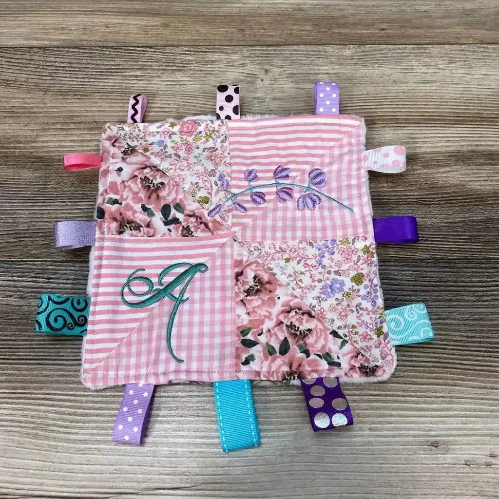 Top-down of pink crinkle square with ribbon tabs showing embroidered initial A, purple vine and multicolored ribbon loops