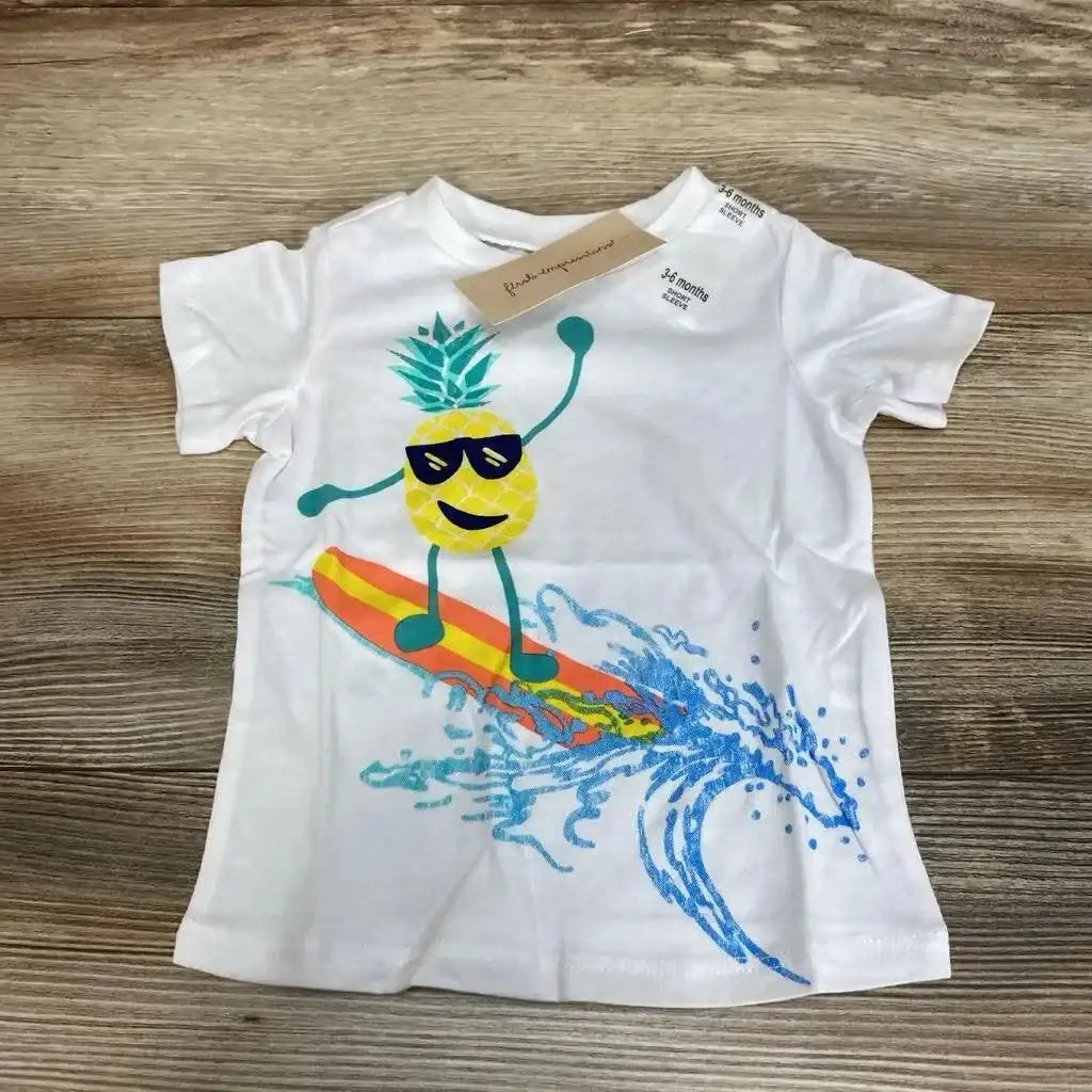 Flat lay of white baby t-shirt (3-6 months) showcasing a surfing pineapple graphic on a wooden tabletop