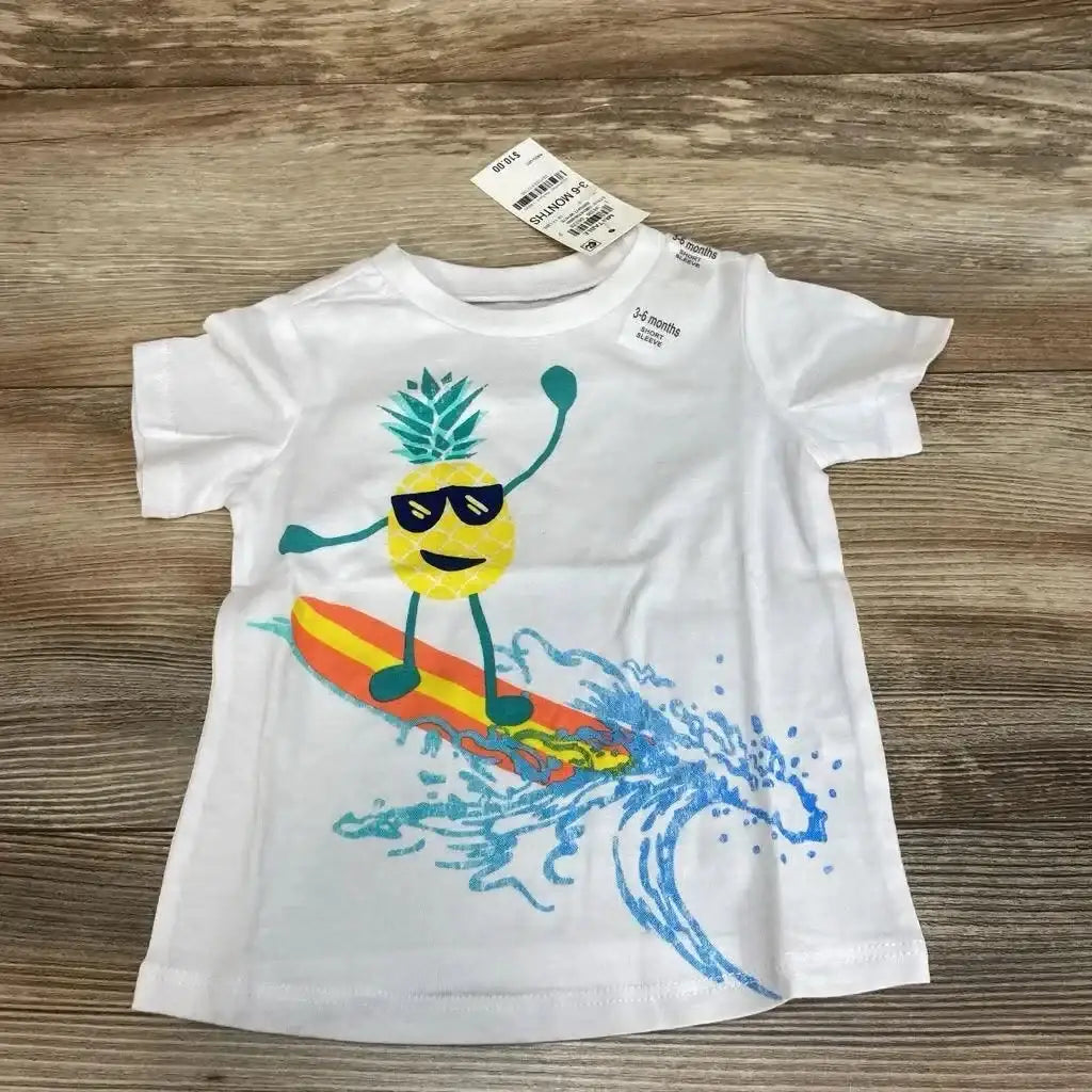 Flat lay of white baby t-shirt showcasing surfing pineapple graphic and visible 3-6 months tag on wood background