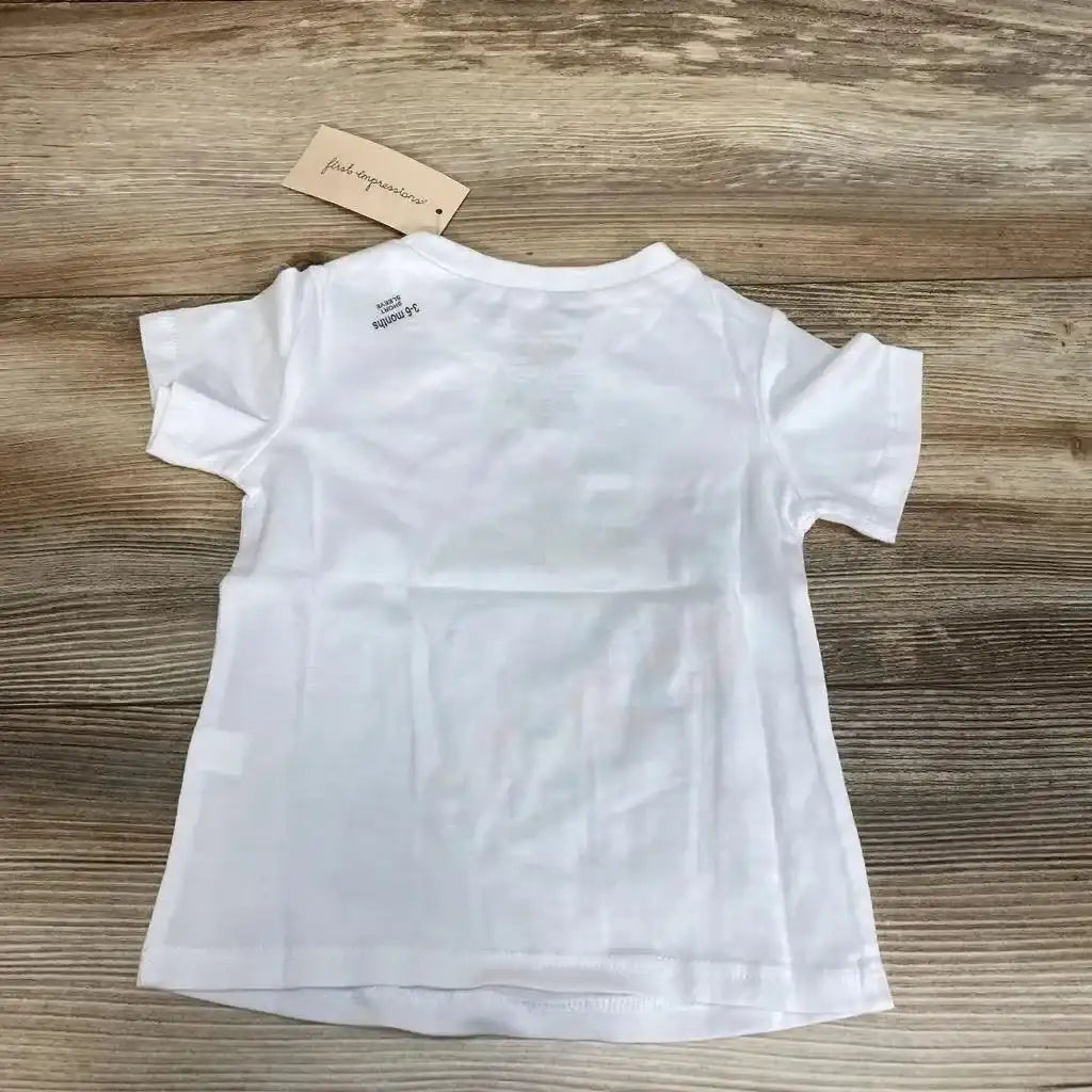 Back view of white First Impressions baby t-shirt (3-6 months) showing short sleeves and attached tag on wood background