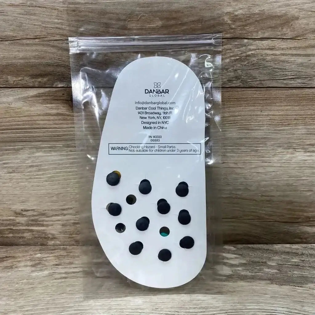 Back view of Danbar Global 10pc clog charms on white shoe-shaped card in clear resealable plastic bag on wooden background