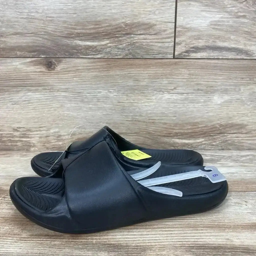 Side view of black Apollo slide sandals showcasing molded textured footbed and visible size tag on wood floor background