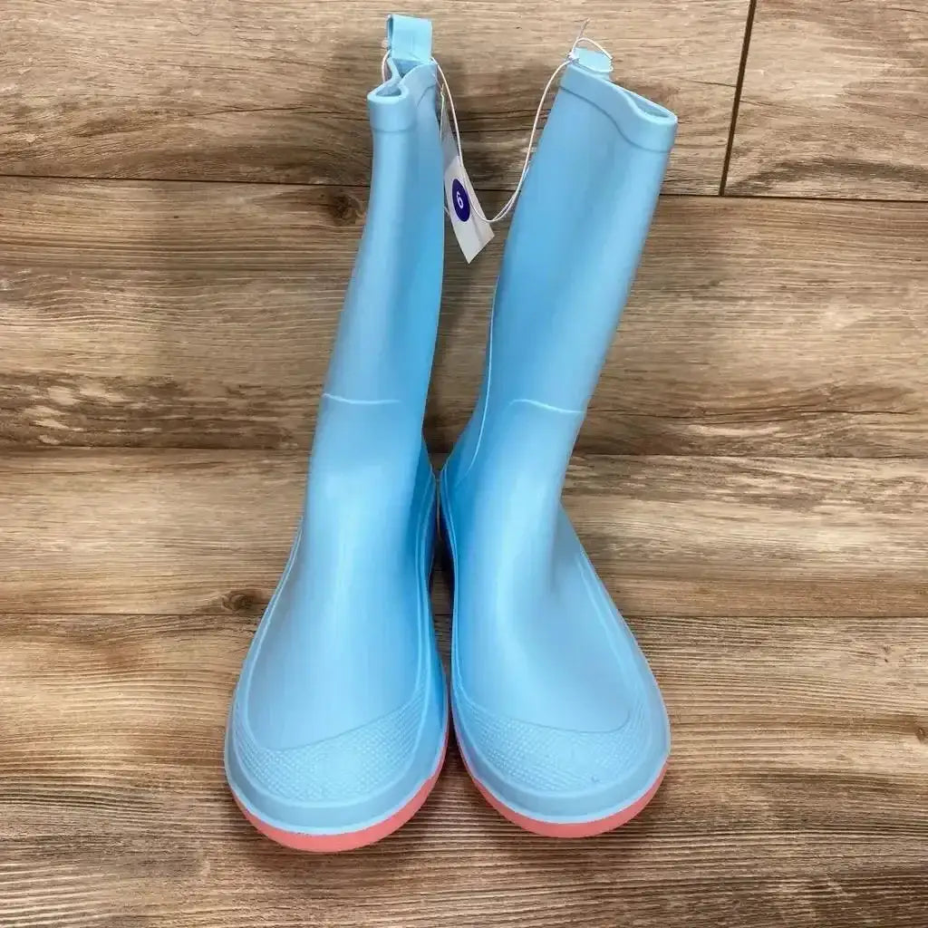 Front view of NEW Cat and Jack Andy Slip-On Rain Boots size 6Y in light blue rubber with pink sole and retail hangtag on wood plank floor