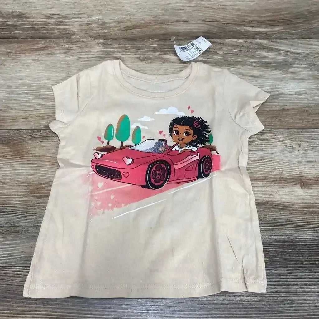 Front flat-lay of beige Children's Place toddler t-shirt showing a girl driving a pink car graphic with hearts and trees on a wood floor