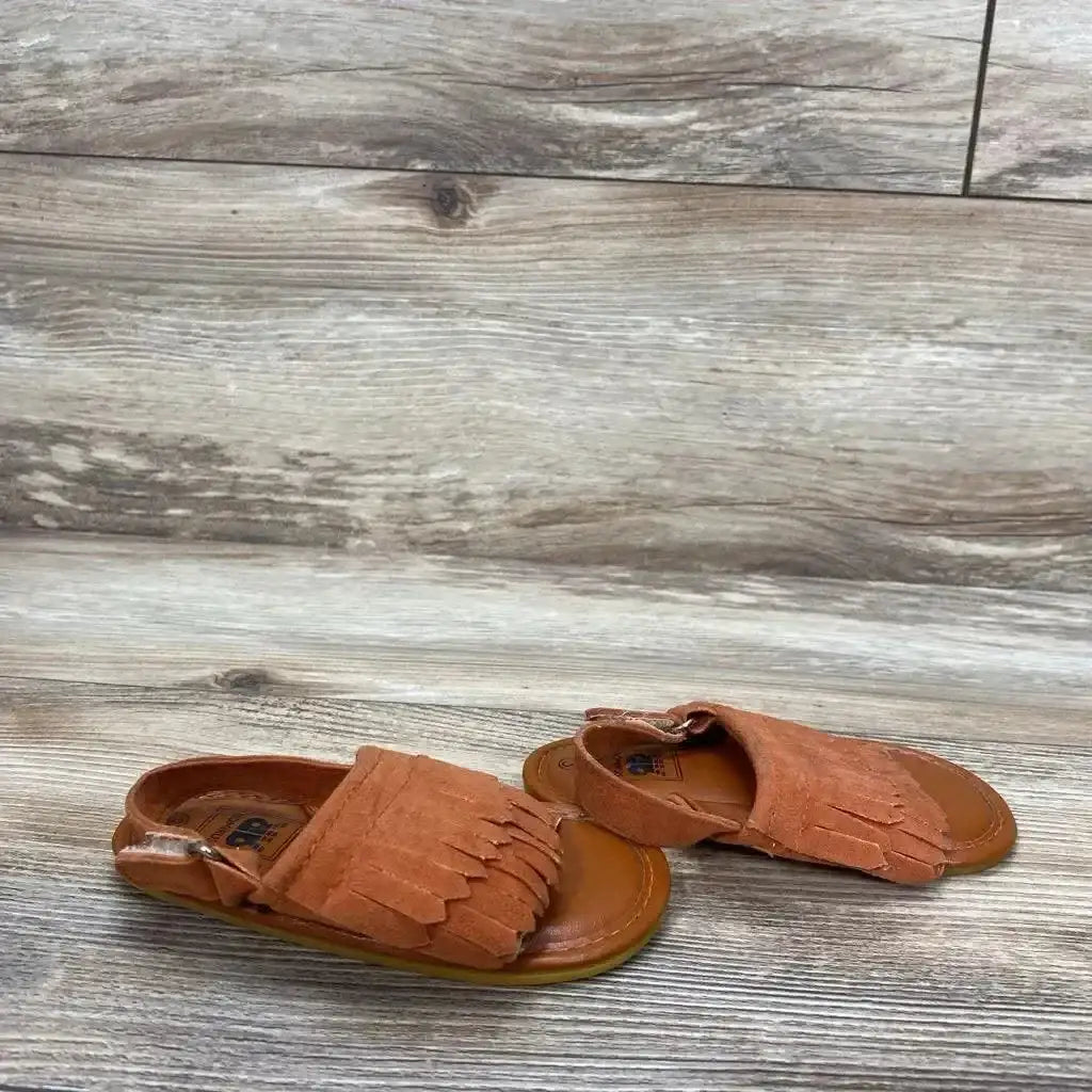 Front-angle view of rust suede toddler moccasins size 3C showing fringe detail and back strap on wood floor