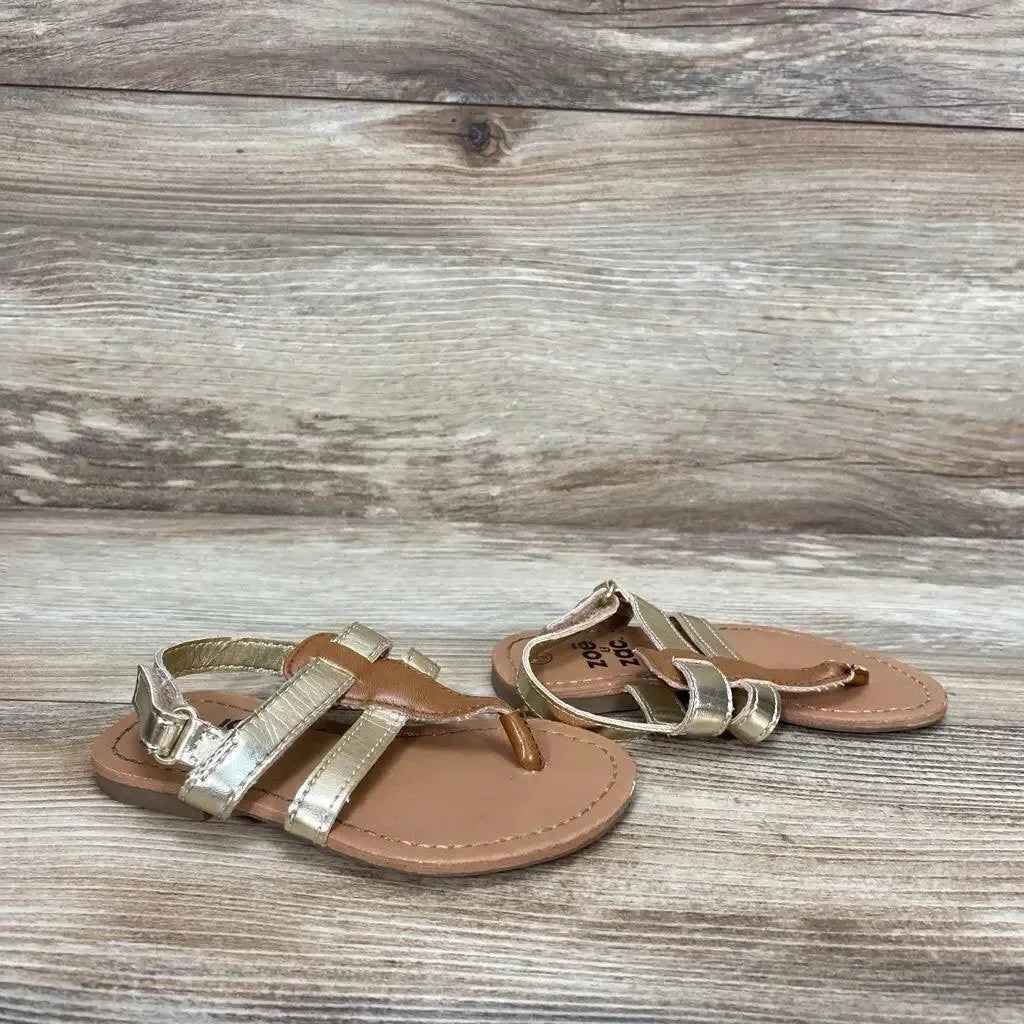 Three-quarter view of Zoe & Zac gold toddler sandals size 6C showing metallic straps and toe-post on wood-look background