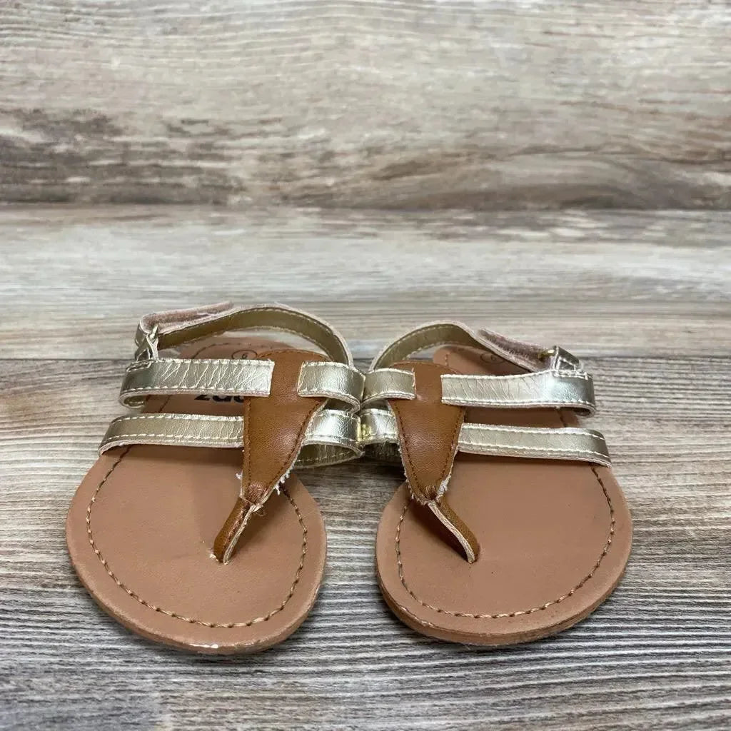 Front view of Zoe & Zac toddler gold sandals with metallic double straps, toe-post and stitched tan footbed on wood background
