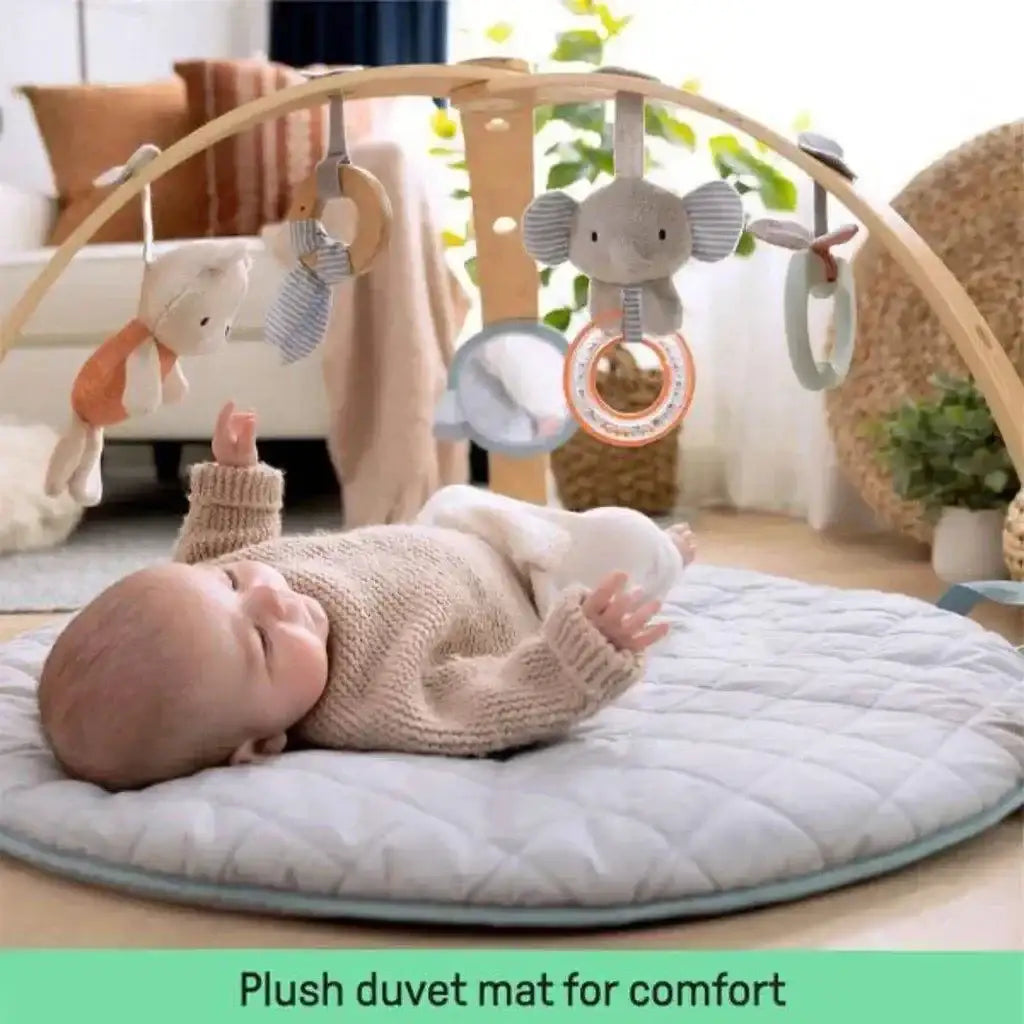 Angled view of Ingenuity Cozy Spot activity gym showing quilted gray mat and wooden arch with hanging toys, baby on mat