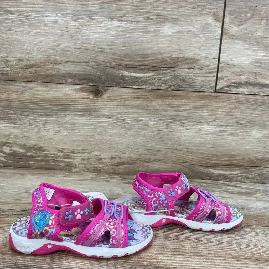 Side view of pink Paw Patrol toddler ankle-strap sandals showcasing paw-print trim, heart applique, and cushioned white sole on wood-look background