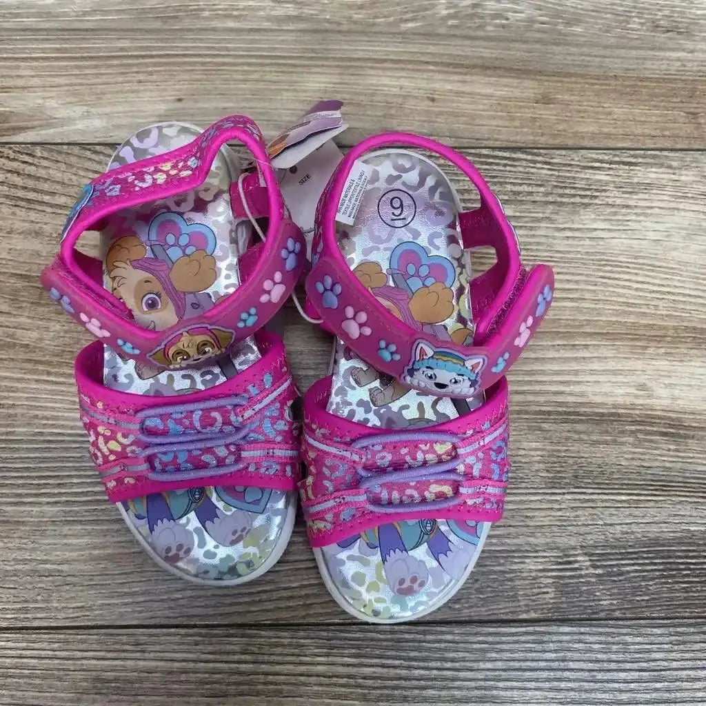Top-down view of pink Paw Patrol ankle-strap sandals, size 9C, showing character insole graphics, paw-print and glitter straps on wood-look floor