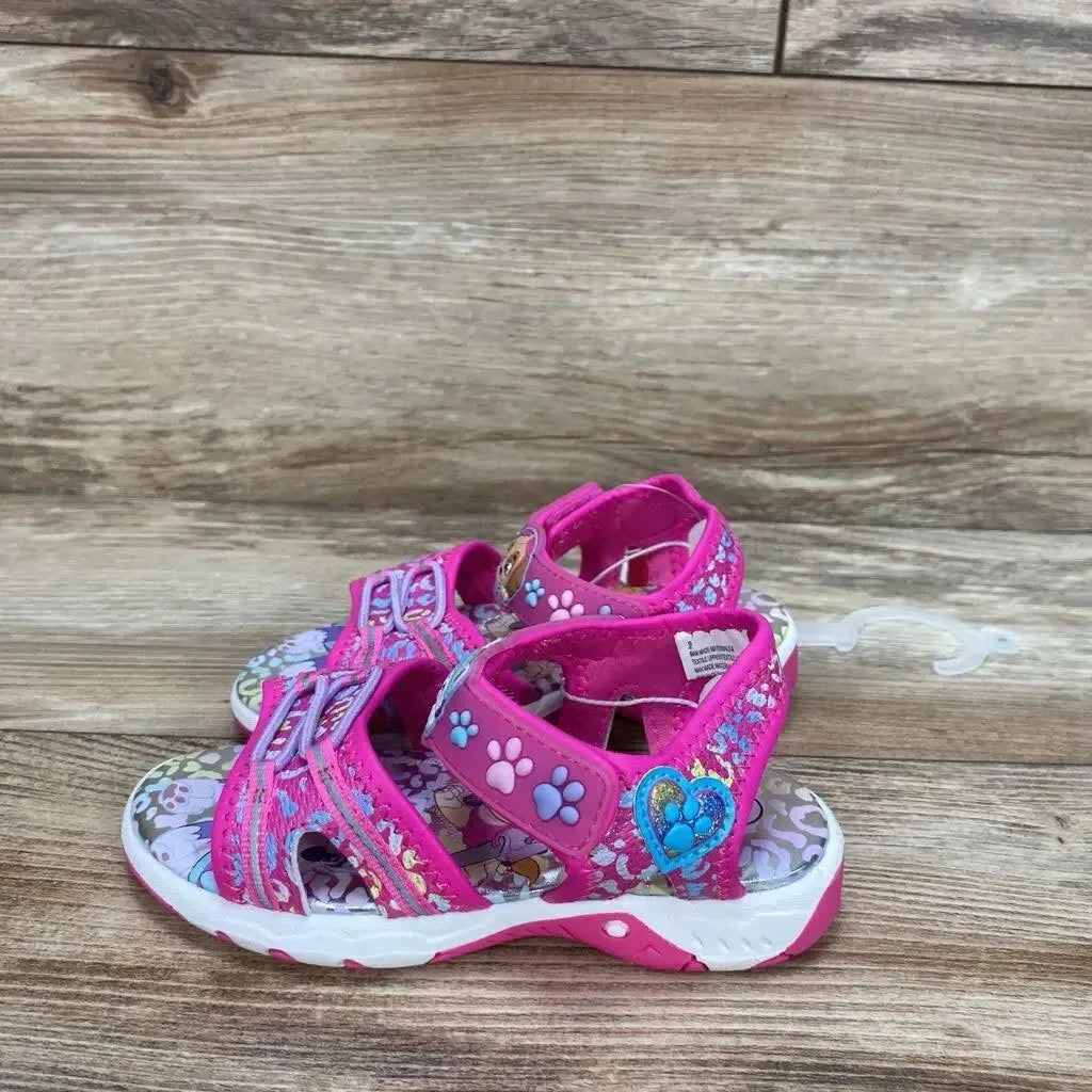 Side view of pink Paw Patrol ankle-strap toddler sandals showing paw-print straps, heart charm, printed character footbed on wood surface