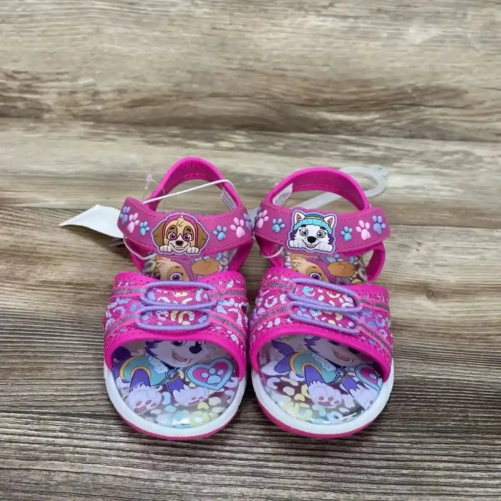 Front view of pink Paw Patrol toddler ankle-strap sandals (size 9C) showing character patches on straps and printed footbeds on wooden surface