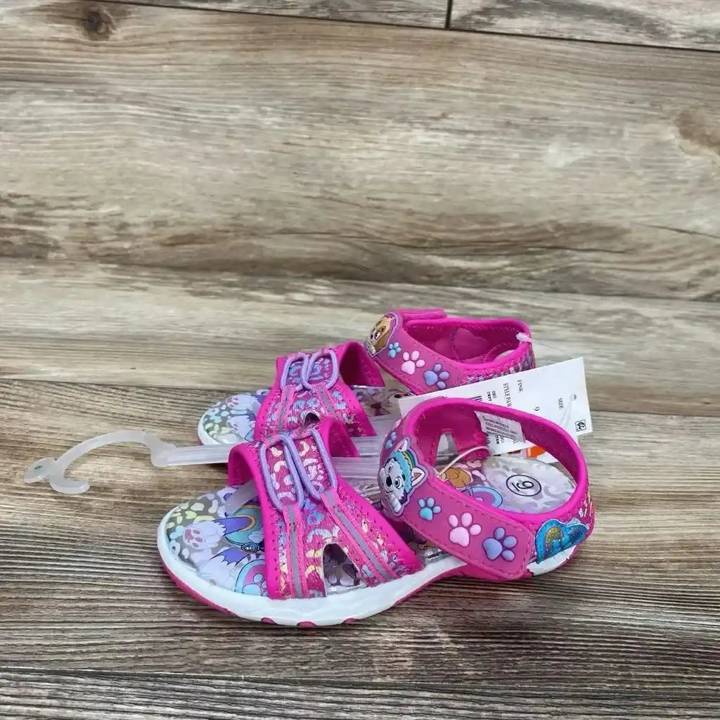 Side view of pink Paw Patrol toddler ankle-strap sandals size 9c showing paw-print straps, character insole, and plastic hanger on wood floor