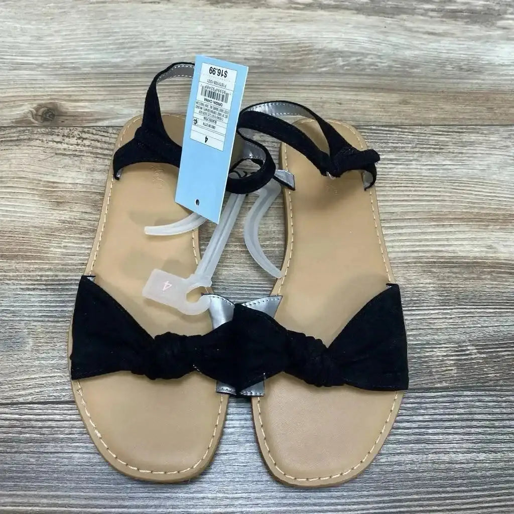 Top-down view of Cat and Jack Lydia girls' sandals showcasing knotted black toe bows and ankle straps on a wood floor background