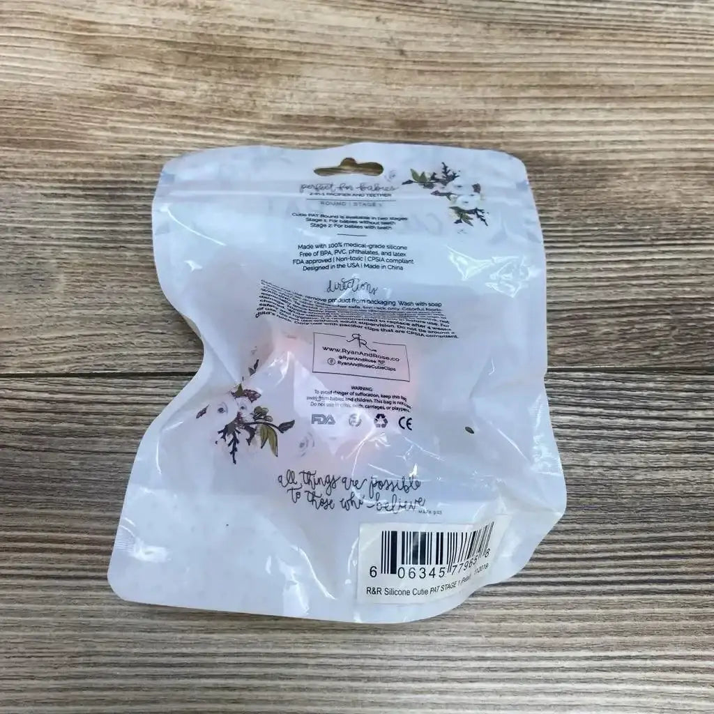 Rear view of Ryan and Rose Cutie PAT round pacifier and teether 0-6m in sealed floral pouch showing barcode on a wooden table