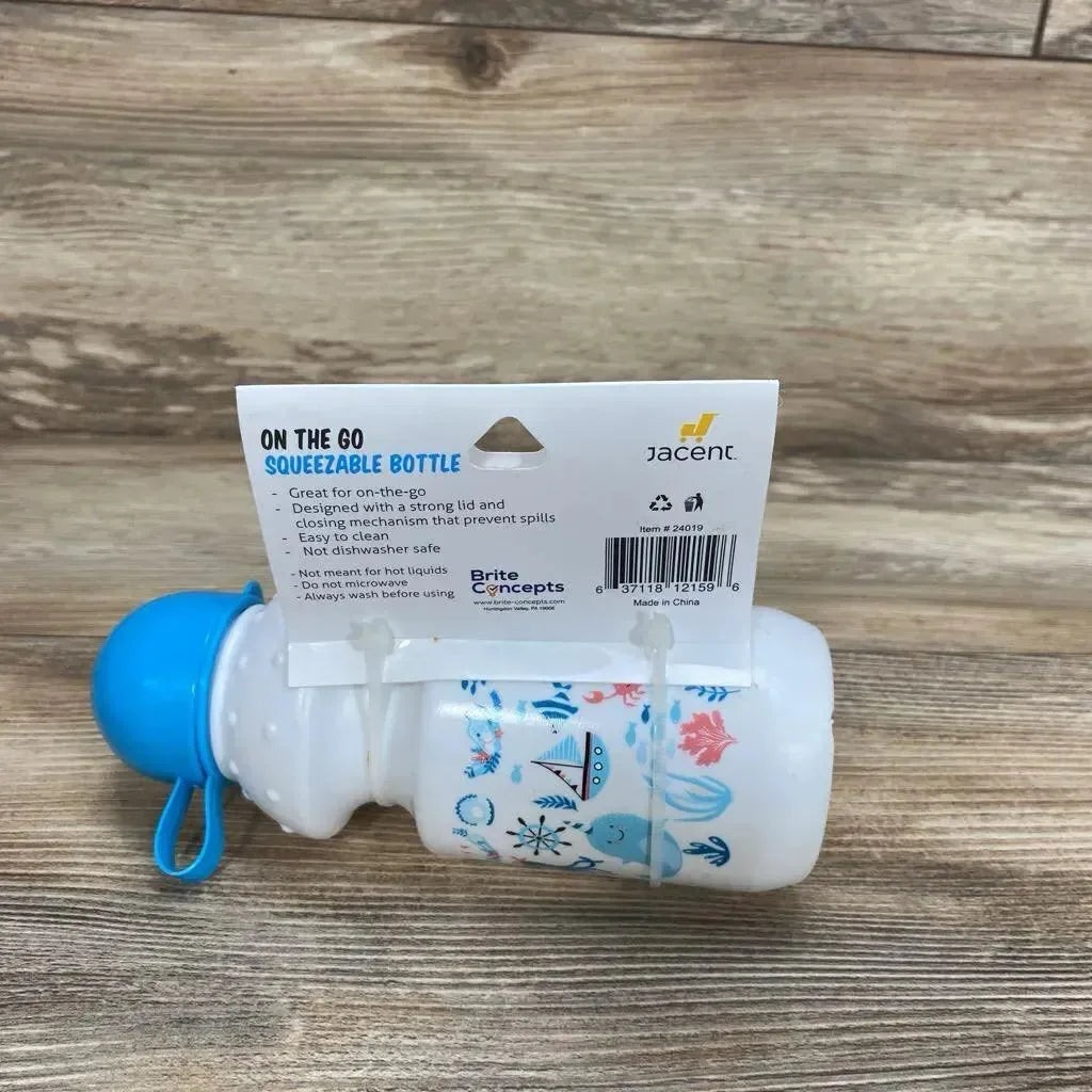 Side view of 13 oz Brite Concepts On The Go squeezable baby bottle with blue cap and nautical prints, attached retail hang card on wood surface