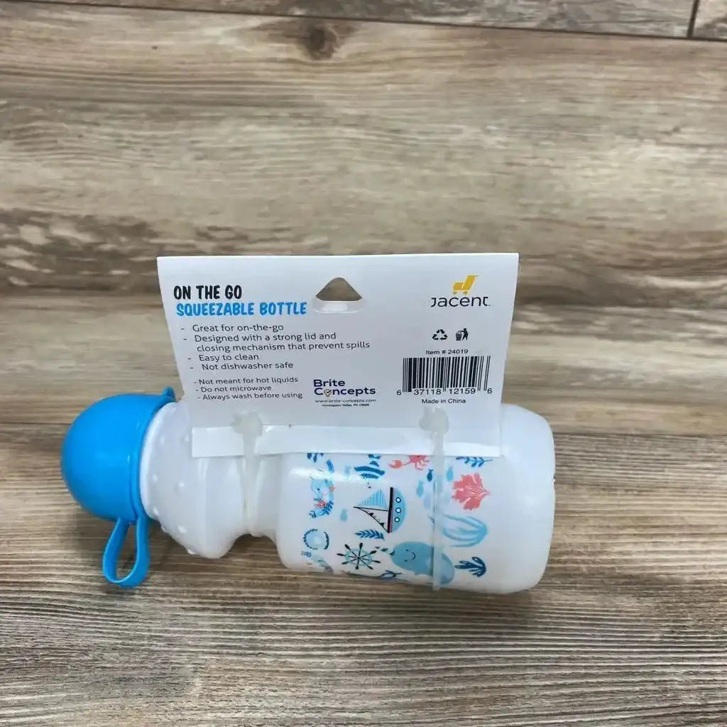 Side view of 13 oz Brite Concepts On The Go squeezable baby bottle with blue cap and nautical prints, attached retail hang card on wood surface