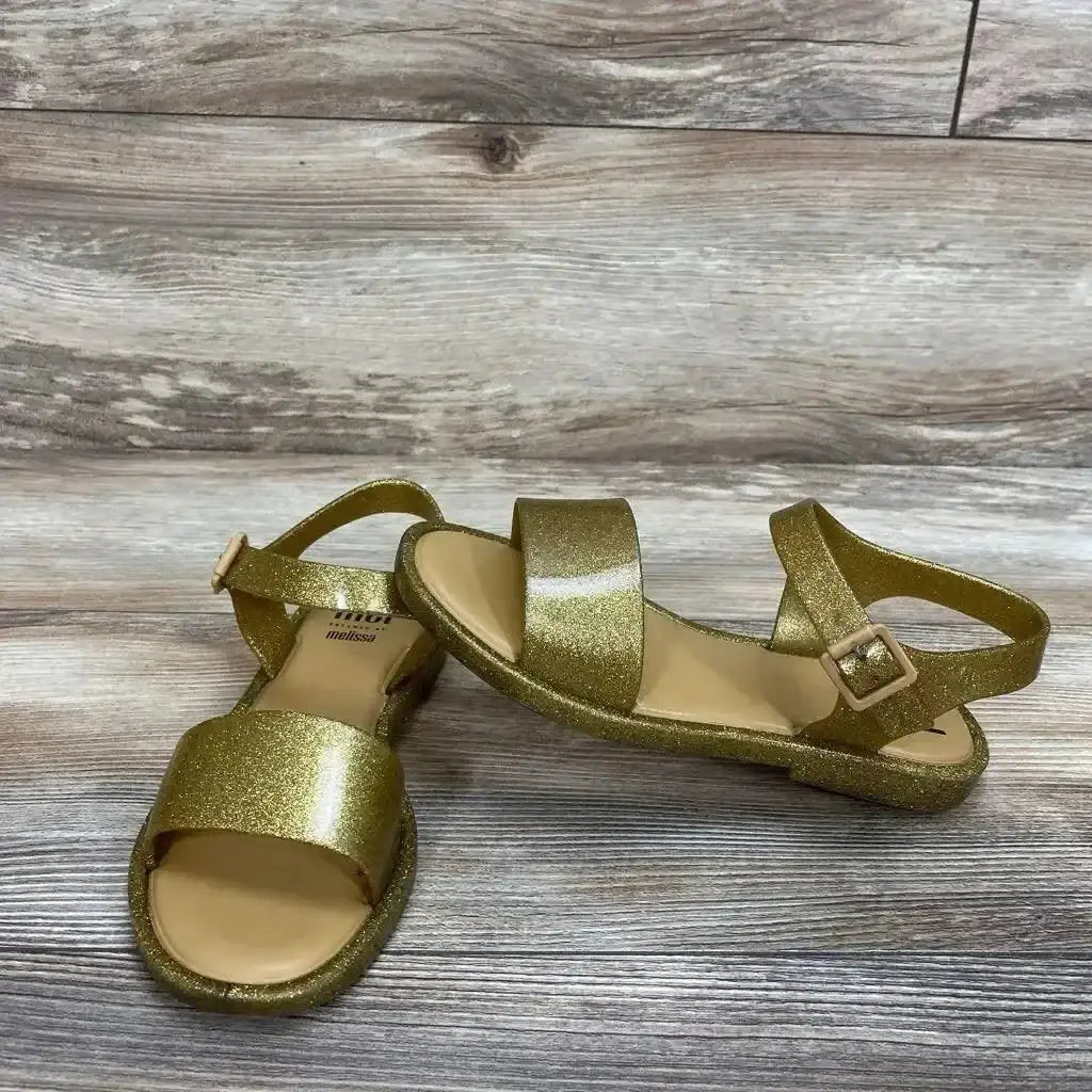 Side view of Mel By Melissa Glitter Jelly Sandals in gold with ankle strap buckle on wood plank floor