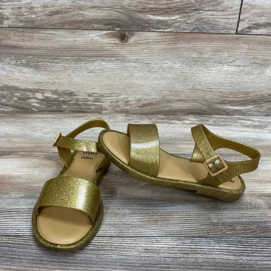Angled view of Mel By Melissa toddler glitter jelly sandals showing gold glitter finish and ankle strap on wood floor