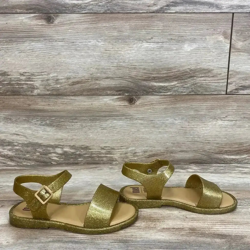Side view of Mel By Melissa gold glitter jelly toddler sandals, ankle strap with buckle and open toe on wood background