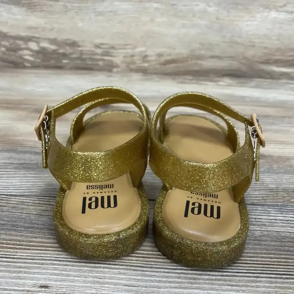 Rear view of toddler Mel by Melissa gold glitter jelly sandals showing ankle straps with buckle on light wood floor