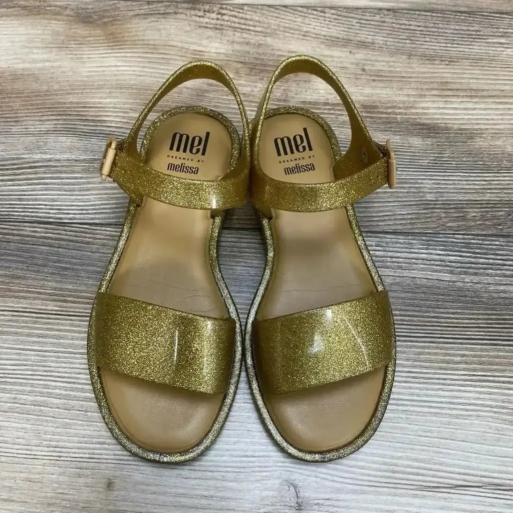 Top view of Mel by Melissa gold glitter jelly sandals, toddler size 2Y, showing ankle strap and wide toe band on wood