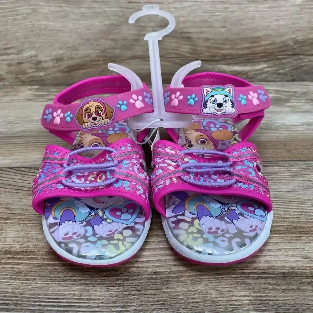 Front view of pink Paw Patrol ankle-strap toddler sandals showcasing character charms, paw-print ankle straps, and printed insoles on wood background