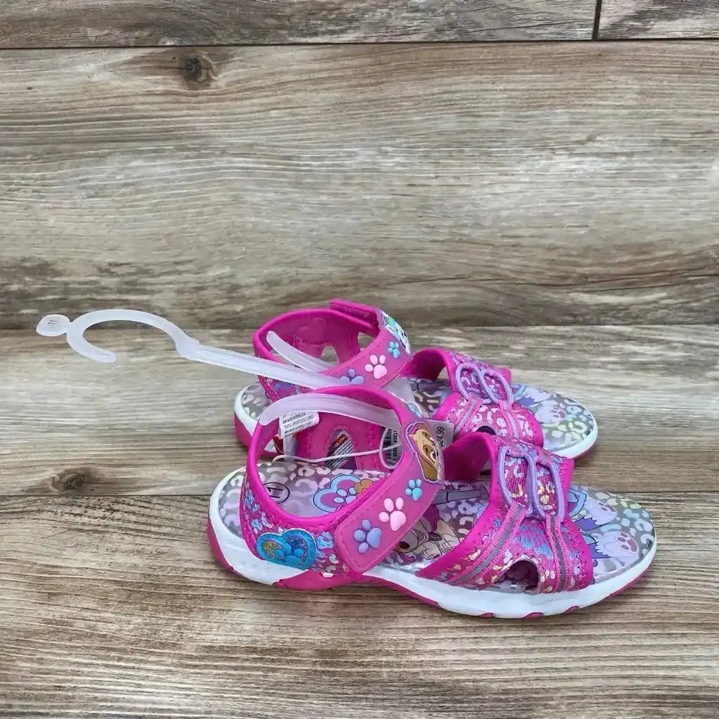 Three-quarter view of pink Paw Patrol ankle-strap toddler sandals showcasing paw-print straps and character insole on wood background, size 11C