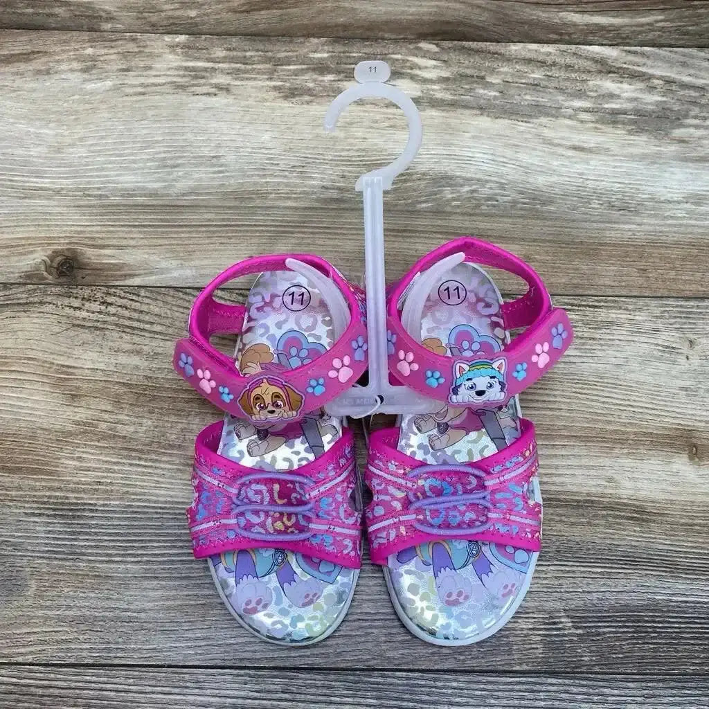 Top-down view of pink Paw Patrol sandals with character charms, paw-print Velcro straps and size 11 tag on wood background