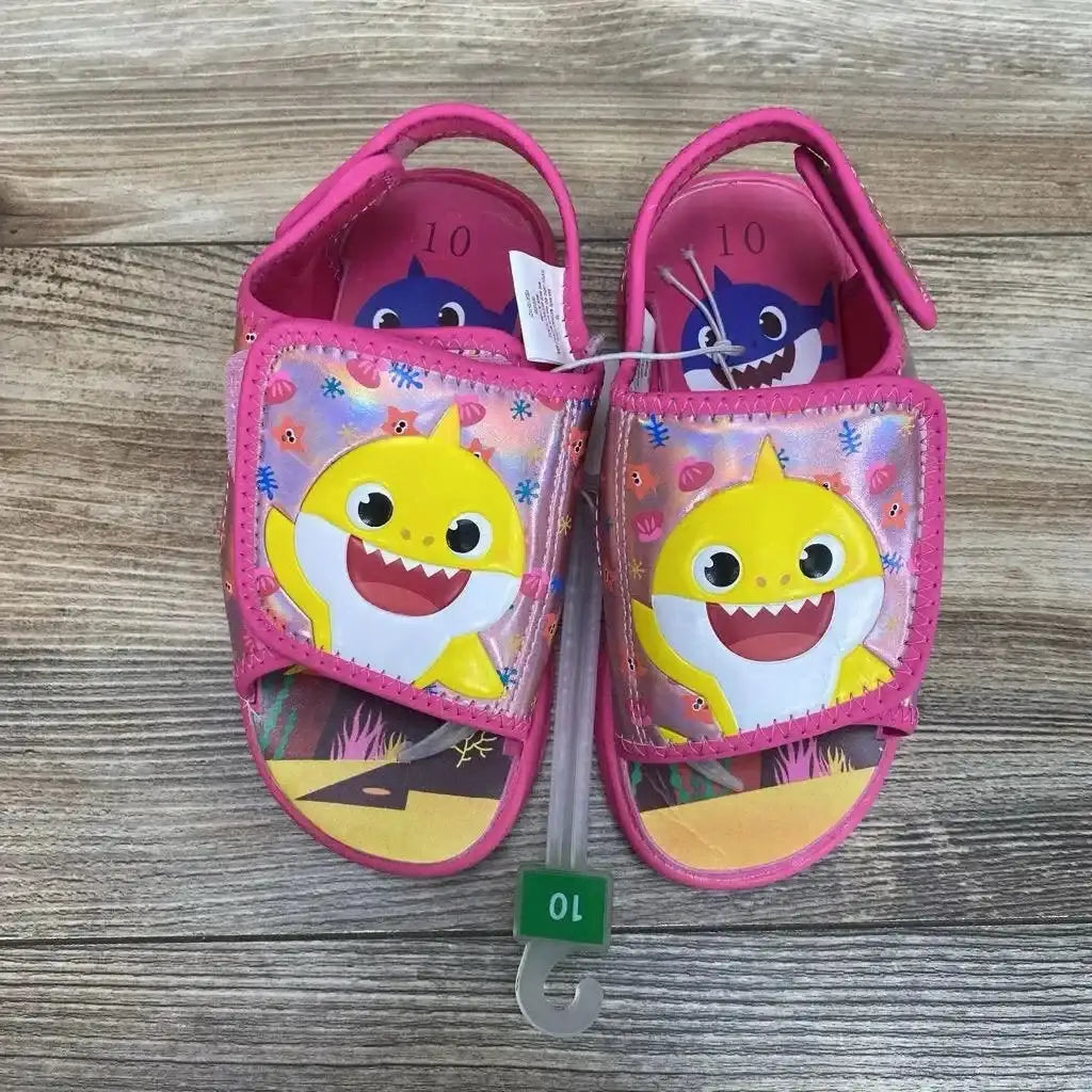 Top view of NEW Pinkfong Baby Shark Water Sandals sz 10c showcasing pink open-toe pair with yellow Baby Shark graphic and size tag on wood plank background