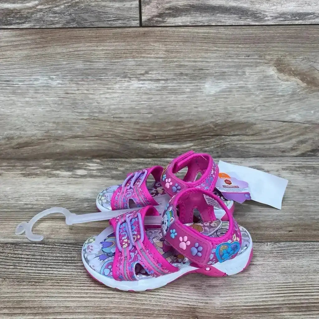 Three-quarter view of pink Paw Patrol ankle-strap toddler sandals showcasing Velcro straps and paw and heart decorations on a wood floor background