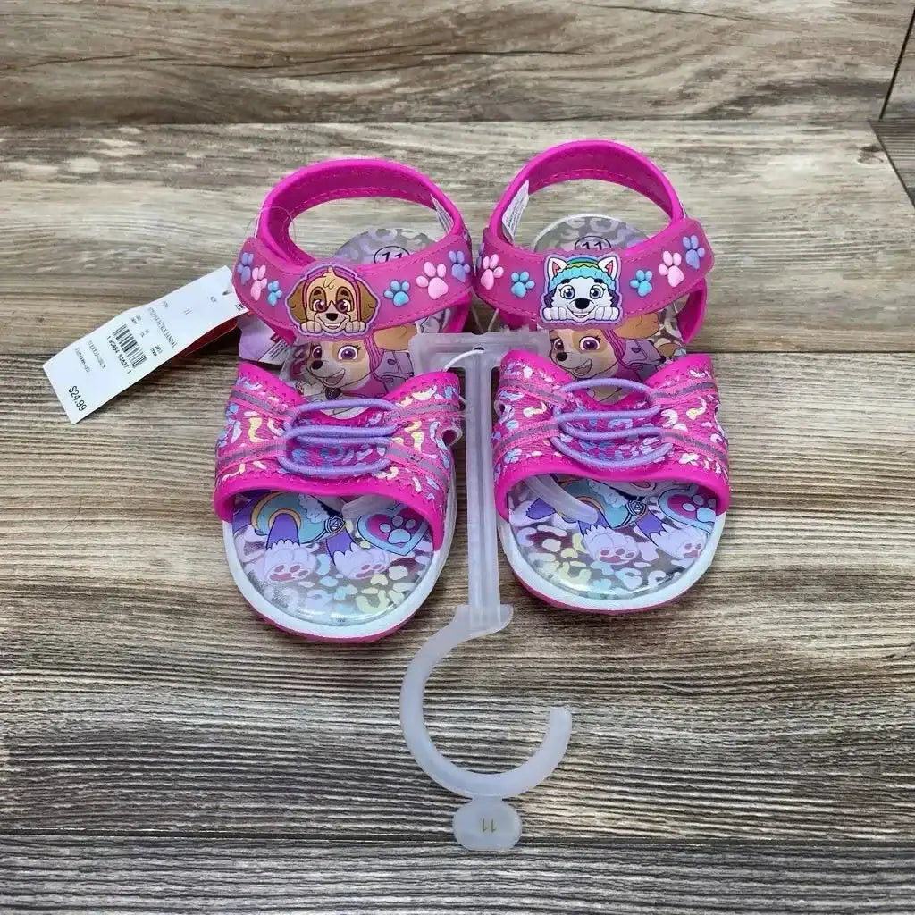 Front view of pink Paw Patrol ankle-strap sandals with character appliqués, glitter velcro straps, size 11C, wood background