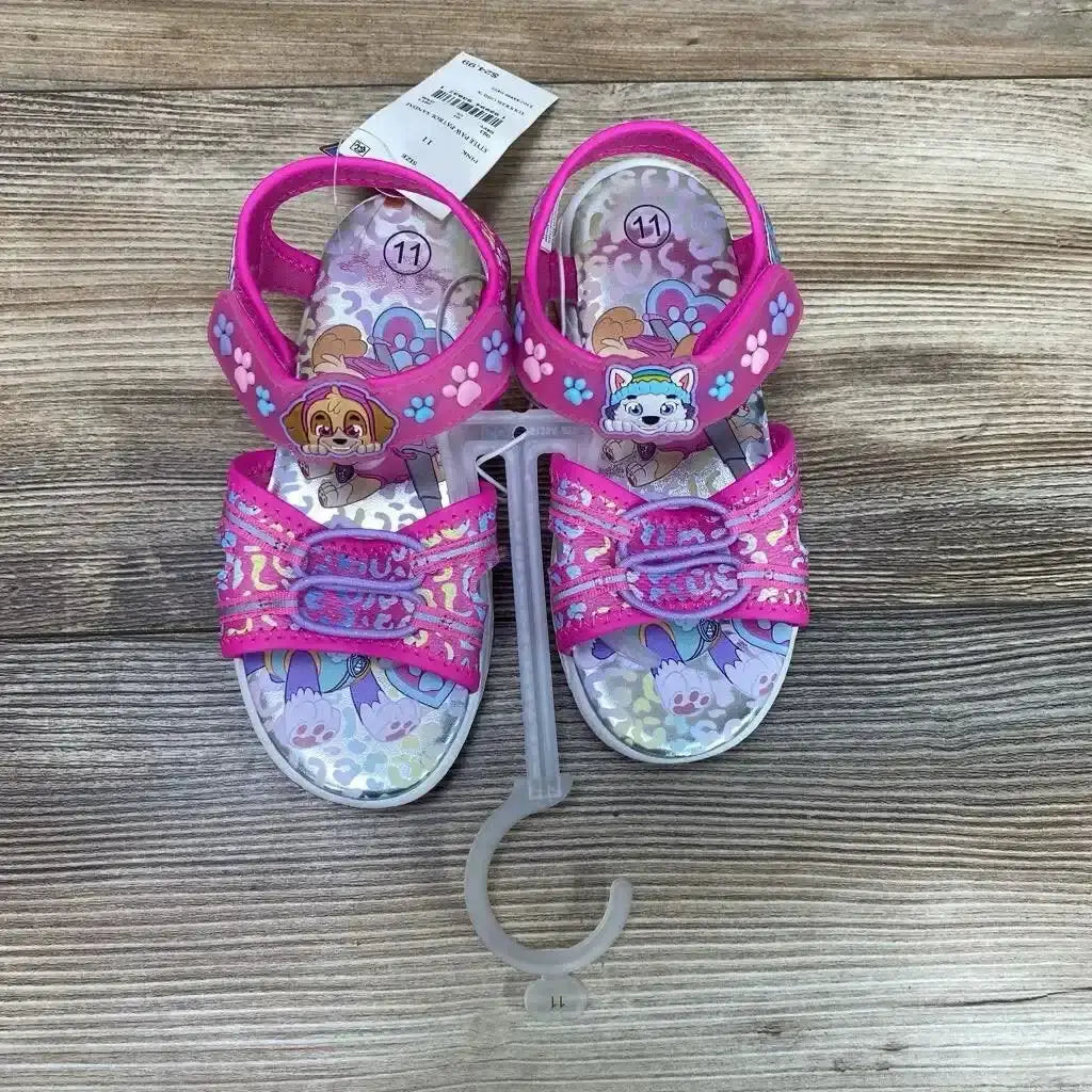 Top-down view of pink Paw Patrol toddler ankle-strap sandals (size 11C) showing character appliques on straps, glittery front straps and printed footbeds on wood background