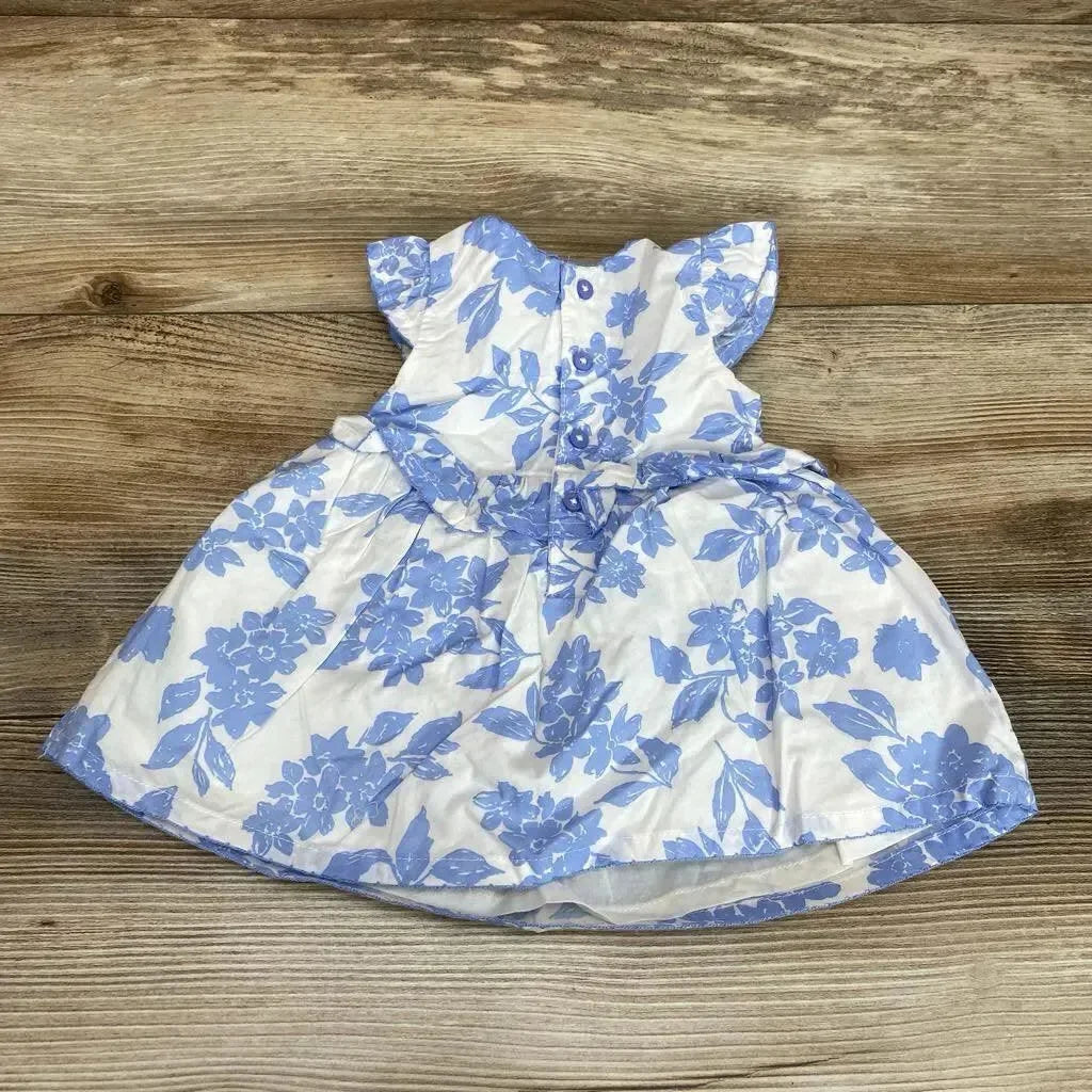 Flat lay front view of newborn white dress showcasing blue floral print, flutter sleeves and back buttons on wood floor background