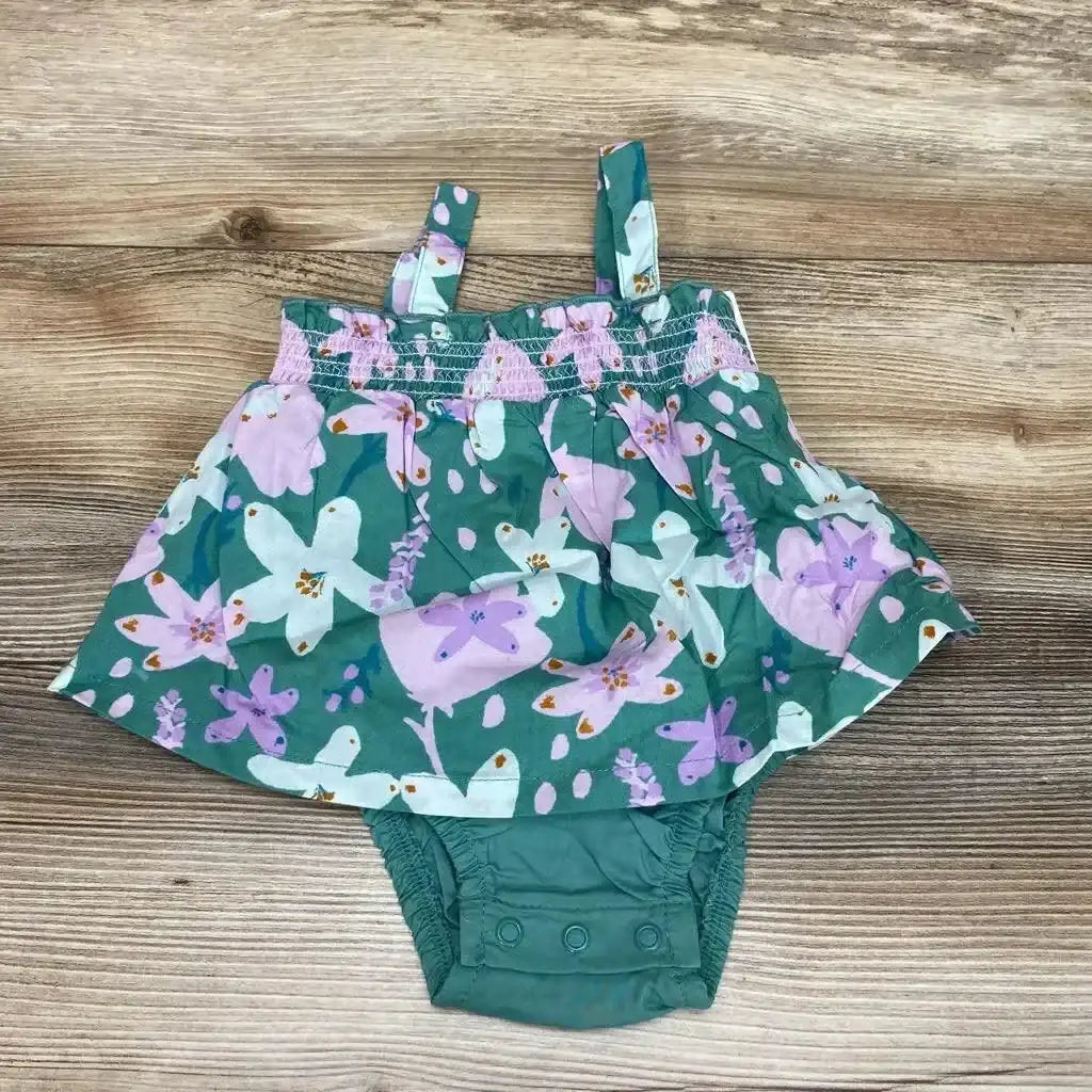 Flat lay of green floral baby bodysuit dress showcasing smocked bodice, shoulder straps, and three snap closures at crotch on wood background