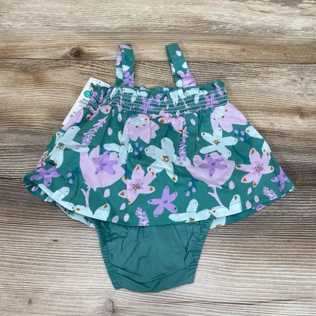 Flat lay of green floral baby bodysuit dress showing smocked top, shoulder straps, and attached bloomers on wood background