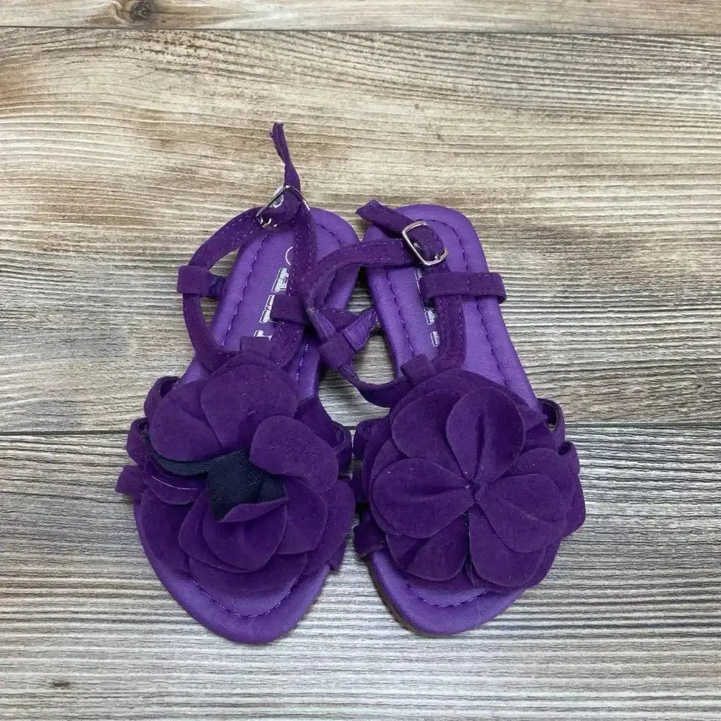 Top-down view of purple toddler strappy sandals showcasing large layered flower on toe and ankle buckle, on wood floor background