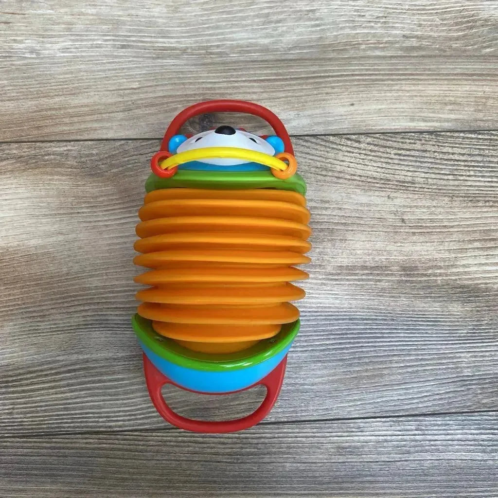 Top-down view of Skip Hop hedgehog accordion toy showcasing orange expandable bellows, red handles, and smiling hedgehog face on wood background